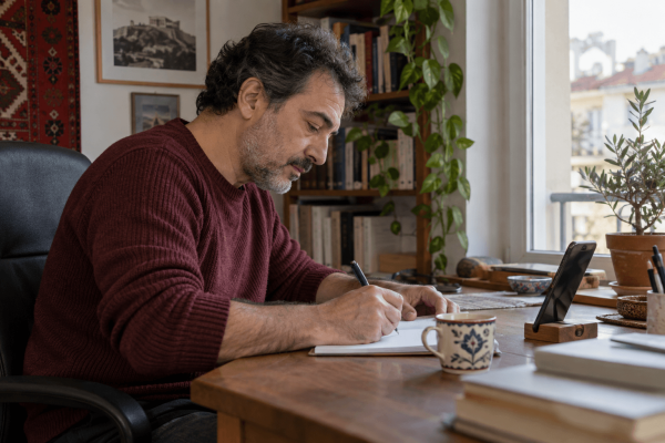 Άνδρας κρατά σημειώσεις σε τετράδιο δίπλα στο κινητό του, που βρίσκεται σε βάση.